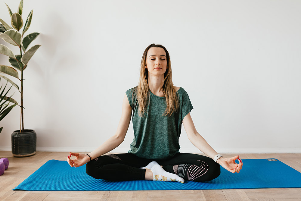 Fizjoterapia oddechowa: Kobieta ubrana w szare spodnie, zieloną koszule i białe skarpety siedzi na niebieskim dywaniku w pozycji lotosa i wykonuje ćwiczenia oddechowe. Z tylu po lewej stronie widnieje kwitek.