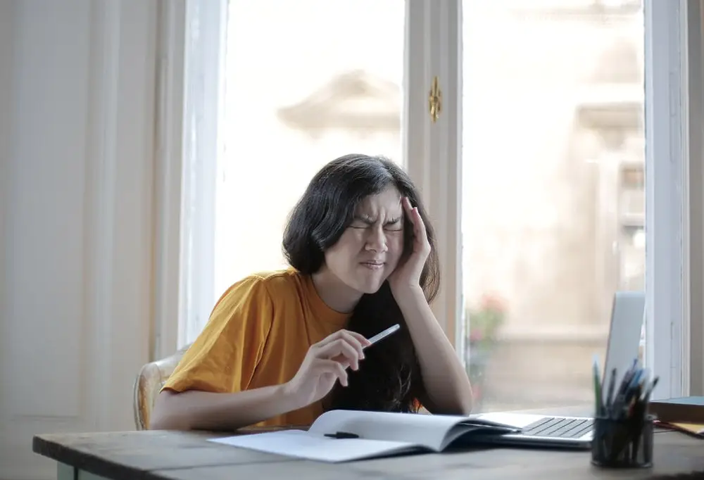 Migraine: The lady in the yellow shirt is sitting behind the desk by the window with her left hand at her head, a pen in her right hand. Her head hurts. There are notebooks on the desk.