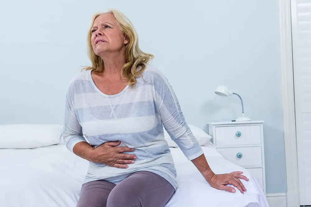 The lady is sitting on the bed, has symptoms of food poisoning, has her hand on her stomach