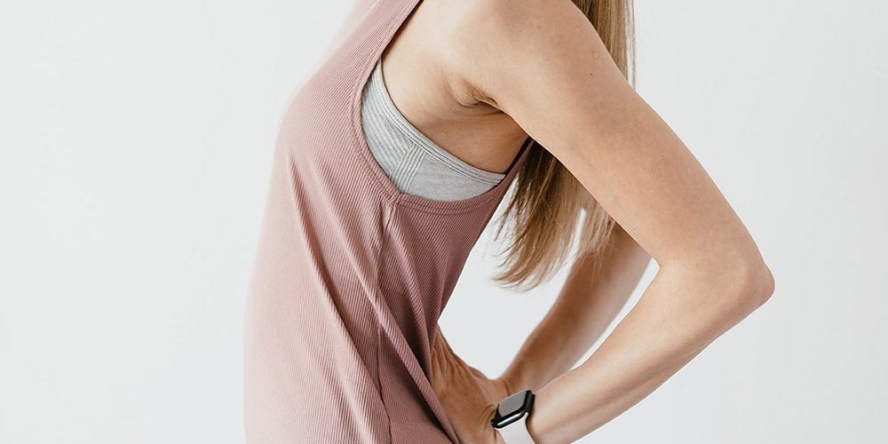 Pani ubrana w beżową podkoszulkę. U Pani boli staw krzyżowo biodrowy i ona trzyma ręce na tym miejscu